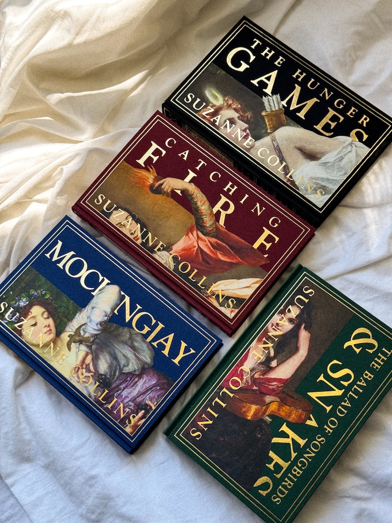 May include: Four hardcover books with gold lettering on the spines. The books are titled "The Hunger Games", "Catching Fire", "Mockingjay", and "The Ballad of Songbirds and Snakes". All four books are by Suzanne Collins.
