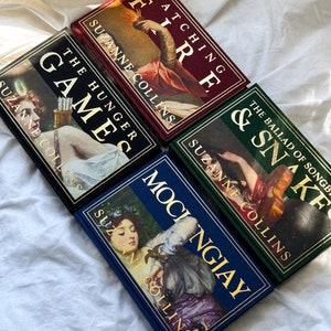 May include: A set of four hardcover books with gold lettering and illustrations on the covers. The books are titled "The Hunger Games", "Catching Fire", "Mockingjay", and "The Ballad of Songbirds and Snakes". All books are by Suzanne Collins.