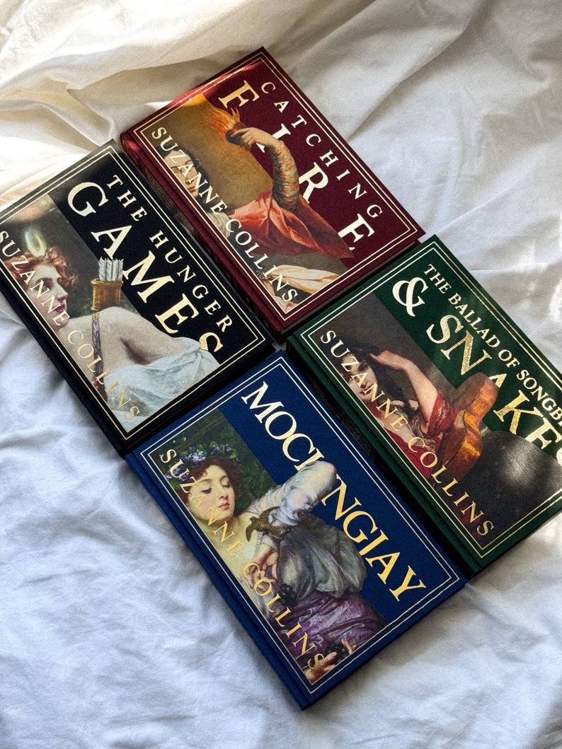 May include: A set of four hardcover books with gold lettering and illustrations on the covers. The books are titled "The Hunger Games", "Catching Fire", "Mockingjay", and "The Ballad of Songbirds and Snakes". All books are by Suzanne Collins.