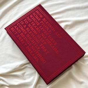 May include: A burgundy book with gold lettering on the cover. The text reads "How blessed are some people, whose lives have no fears, no dreads; to whom sleep is a blessing that comes nightly, and brings nothing but sweet dreams."