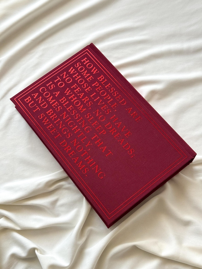 May include: A burgundy book with gold lettering on the cover. The text reads "How blessed are some people, whose lives have no fears, no dreads; to whom sleep is a blessing that comes nightly, and brings nothing but sweet dreams."