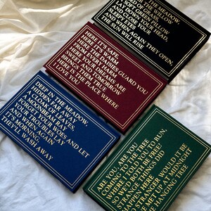 May include: Four small hardcover books with gold lettering on a white background. The books have different colored covers: black, burgundy, navy blue, and green. The text on the covers reads: "Deep in the meadow, under the willow, a bed of grass, a soft green pillow, lay down your head, and close your sleepy eyes, and when again they open, the sun will rise." "Here it's safe, here it's warm, here the daisies guard you from every harm, here your dreams are sweet and tomorrow brings them true, here is the place where I love you." "Deep in the meadow, hidden far away, a cloak of leaves, a moonbeam ray, forget your woes and let your troubles lay, and when again it's morning, they'll wash away." "Are you, are you coming to the tree where I told you to run, so we'd both be free? Strange things did happen there, no stranger would it be if we met up at midnight in the hanging tree."