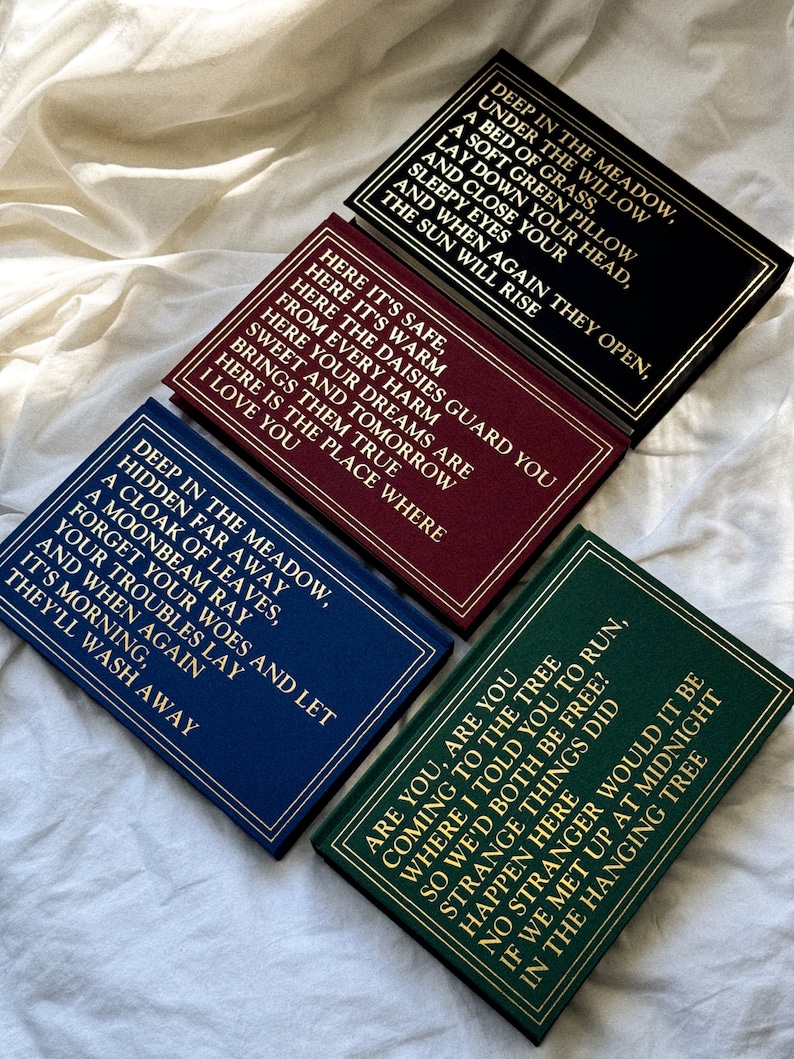 May include: Four small hardcover books with gold lettering on a white background. The books have different colored covers: black, burgundy, navy blue, and green. The text on the covers reads: "Deep in the meadow, under the willow, a bed of grass, a soft green pillow, lay down your head, and close your sleepy eyes, and when again they open, the sun will rise." "Here it's safe, here it's warm, here the daisies guard you from every harm, here your dreams are sweet and tomorrow brings them true, here is the place where I love you." "Deep in the meadow, hidden far away, a cloak of leaves, a moonbeam ray, forget your woes and let your troubles lay, and when again it's morning, they'll wash away." "Are you, are you coming to the tree where I told you to run, so we'd both be free? Strange things did happen there, no stranger would it be if we met up at midnight in the hanging tree."
