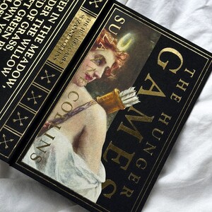 May include: A black book with gold lettering titled "The Hunger Games" by Suzanne Collins. The cover features a woman with an arrow and a bow.