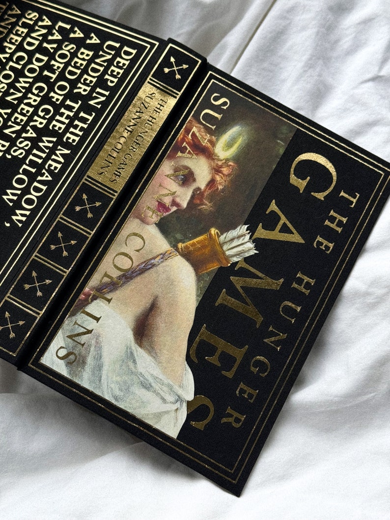 May include: A black book with gold lettering titled "The Hunger Games" by Suzanne Collins. The cover features a woman with an arrow and a bow.