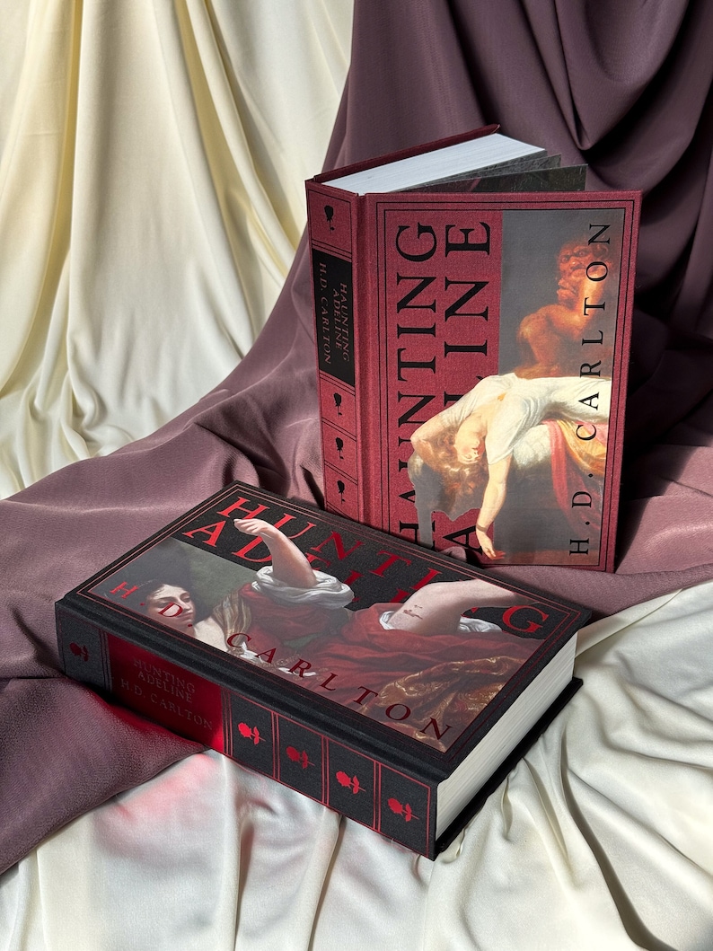 Puede incluir: Dos libros de tapa dura con cubiertas negras y lomos rojos. Los libros se titulan "Hunting Adeline" y "Hunting Adeline". Las cubiertas presentan pinturas de mujeres con vestidos rojos.