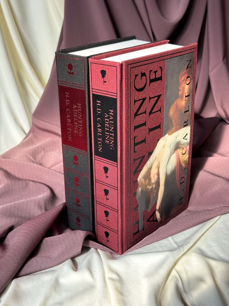Puede incluir: Dos libros de tapa dura con cubiertas rojas y negras. El libro de la derecha tiene el t&iacute;tulo "HAUNTING ADELINE" y el autor "H.D. CARLTON". El libro de la izquierda tiene el t&iacute;tulo "HUNTING ADELINE" y el autor "H.D. CARLTON".