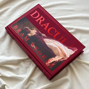 May include: A red hardcover book titled "Dracula" by Bram Stoker. The cover features a painting of a woman in a white dress lying on a bed.