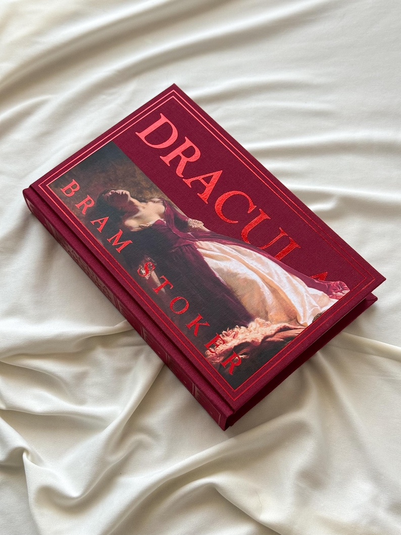 May include: A red hardcover book titled "Dracula" by Bram Stoker. The cover features a painting of a woman in a white dress lying on a bed.