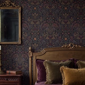 Op de afbeelding: Een slaapkamer met donker bloemenbehang, een goudkleurige spiegel en een houten nachtkastje met een koperen kandelaar. Een bed met een gouden hoofdeinde is opgemaakt met een dieppaars fluwelen dekbed en bijpassende fluwelen kussens.
