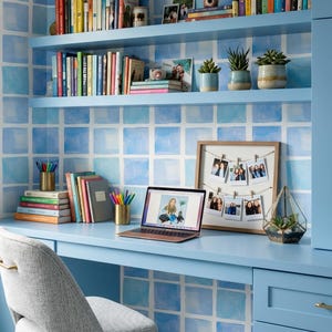 Puede incluir: Una oficina en casa con un escritorio, estanterías y una pared azul claro. Las estanterías contienen libros, plantas y fotos. Un ordenador portátil está sobre el escritorio, junto con cuadernos, bolígrafos y una exhibición de fotos enmarcadas. Una silla gris completa el espacio de trabajo.