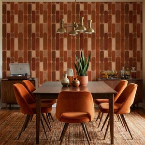 a modern dining room with a wooden table surrounded by orange upholstered chairs. The walls are covered in a patterned wallpaper, and a chandelier hangs above the table. The room also features a record player and a potted plant, adding to the vintage-inspired ambiance.