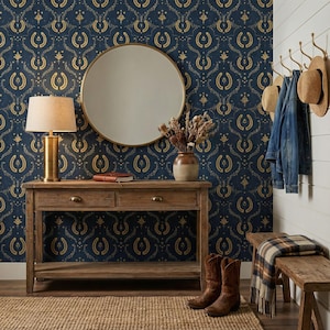 a stylish entryway with a wooden console table, a round mirror, and a wooden bench. The wall behind the console table has a blue and gold patterned wallpaper. On the table, there is a lamp, a vase with flowers, and a pair of boots. The overall scene has a rustic and elegant aesthetic.
