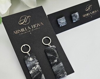 May include: A pair of black and silver dangle earrings with silver hoops and rectangular black and silver resin pieces. The earrings are displayed on a black card with the text "Armilla Nova" in gold lettering.