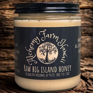 May include: A jar of raw Big Island honey from Alchemy Farm Hawaii. The jar has a black lid and a dark blue label with white text and a tree logo. The honey is a light beige color and the jar is sitting on a wooden surface.