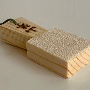 May include: A wooden sanding block with a rough, white surface and a handle with Japanese characters. The block is made of light wood and has a green string attached to the handle.
