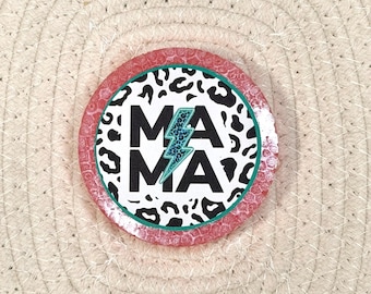 May include: A circular pin featuring a white background with a black leopard print border. The word MAMA is printed in bold black letters, with a teal leopard print lightning bolt replacing the letter A in the center of the design.