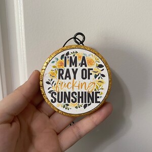 May include: A round, gold-rimmed ornament with a white background and floral design. The ornament features the text "I'm a ray of fucking sunshine".
