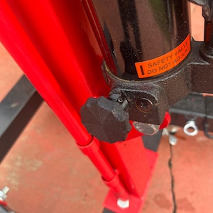 May include: Close-up of a red and black hydraulic lift with a black knob and a safety valve sticker that reads "Safety Valve Do Not Adjust".
