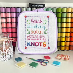 Könnte beinhalten: Ein lilafarbener Stickrahmen mit dem Text "brick couching satin long & short shading KNOTS" in einem farbenfrohen Blumendesign. Der Rahmen ist von Garnrollen in verschiedenen Farben umgeben.