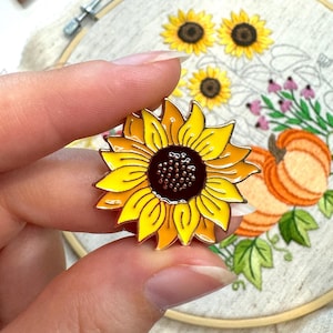May include: A sunflower enamel pin in yellow and brown with a gold border. The pin is held in front of an embroidery hoop with a sunflower design. The background includes a small pin cushion and embroidery threads.
