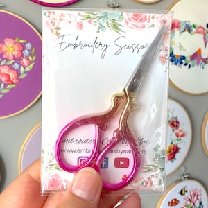 May include: Embroidery scissors with a gold-coloured handle and bright pink finger holes, packaged in a clear plastic sleeve. The packaging has floral accents and the words "Embroidery Scissors". Several embroidered hoops are in the background.