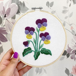 May include: Embroidered floral hoop art featuring purple and yellow pansies with green leaves and stems. The embroidery is set against a white fabric within a wooden hoop. The background is a floral wallpaper.