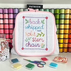 May include: A pink embroidery hoop with white fabric inside. The fabric has colorful embroidery that reads "back whipped back split stem chain". The hoop is surrounded by spools of thread in various colors.