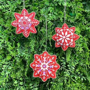 May include: Three red snowflake ornaments with silver trim and white and purple embellishments hang against a green leafy background. The ornaments are star-shaped and appear to be handmade, perfect for holiday decor.