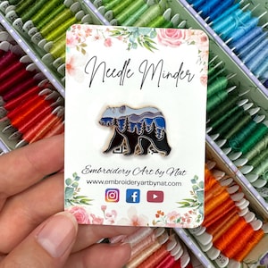 May include: A needle minder with a bear design, featuring a black bear silhouette with a mountain and forest scene in shades of blue and grey. The card reads "Needle Minder" and "Embroidery Art by Nat". The background shows spools of colourful embroidery floss.