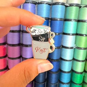 May include: An enamel pin depicting three stacked teacups, with the words "Sip" and "Knit". The pin has a gold border and is set against a backdrop of multi-coloured thread spools.