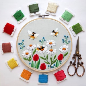 May include: A close-up of a hand-stitched embroidery hoop with a floral design featuring bees and flowers. The hoop is surrounded by colorful embroidery floss spools.