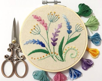 May include: A colourful embroidery hoop with a floral design featuring lavender, green leaves, and yellow accents. The hoop is surrounded by colourful tassels.