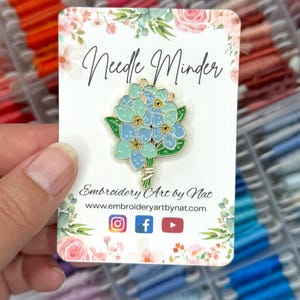 May include: A floral needle minder pin featuring a bouquet of blue and teal flowers with green leaves. The pin is on a card with the text "Needle Minder" and "Embroidery Art by Nat". The card has a floral design.
