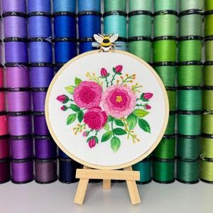 Peut inclure: Un cercle de broderie blanc avec un motif floral de roses roses et de feuilles vertes. Le cercle est sur un support en bois et est entouré de bobines de fil coloré.