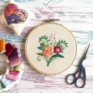 May include: A close-up of a white embroidery hoop with a floral design embroidered in pink, orange, and green thread. The hoop is on a light blue and white wooden surface. A pair of rainbow-coloured embroidery scissors are also in the image.