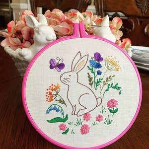 May include: Embroidered hoop art featuring a white bunny sitting in a field of colorful flowers. The bunny is surrounded by pink, purple, blue, yellow, and green flowers. The hoop is pink.