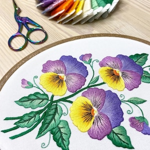 May include: Embroidered pansies in a wooden hoop. The flowers are yellow and purple with green leaves. Embroidery floss in various colors and iridescent scissors are in the background. A craft project.