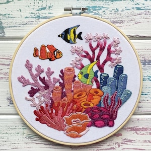May include: Embroidered hoop art featuring an underwater scene with colorful coral and three fish. The fish are orange, yellow, blue, and black. The coral is various shades of pink, orange, and blue. The embroidery is on a white background.