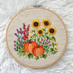 May include: Embroidered hoop art featuring a autumnal scene with sunflowers, lavender, and pumpkins. The hoop is made of wood and has a beige fabric background.