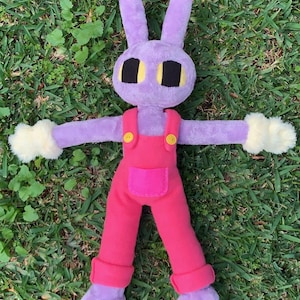 Op de afbeelding: Een pluche konijn met een paars lichaam, lange oren en zwarte ogen met gele accenten. Het draagt een roze overall met gele knopen en crèmekleurige, donzige handschoenen. Het speelgoed ligt op groen gras.