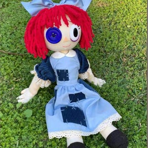 May include: A rag doll with bright red yarn hair, a light blue satin hair bow, and a mismatched eye design featuring one large blue button and one white eye. The doll wears a light blue dress with dark blue corduroy patches and white lace trim.
