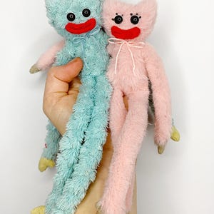 Może przedstawiać: Dwie pluszowe zabawki, jedna jasnoniebieska i jedna różowa, trzymane są w dłoni. Każda ma uśmiechniętą twarz z czarnymi oczami i czerwonymi ustami. Zabawki mają długie, puszyste kończyny i żółte stopy. Niebieska zabawka ma niebieską kokardę, a różowa zabawka ma różową kokardę.