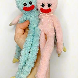 Op de afbeelding: Twee pluche poppen met lange, pluizige ledematen en lichamen. De ene pop is lichtblauw, de andere lichtroze. Beide hebben ronde zwarte knoopogen, brede rode gestikte monden en kleine strikjes bij de nek. De handen en voeten zijn beige van kleur.