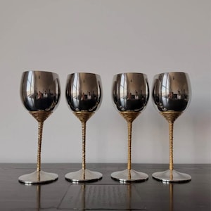 May include: Four wine glasses with silver-colored bowls and bases. The stems are gold-colored and have a twisted rope design. The glasses are arranged in a row on a dark wooden surface.