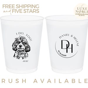May include: Two white plastic cups with black text and designs. One cup says "I do, too!" with a black and white illustration of a dog. The other cup says "Daniel & Helene" with their initials in a circle and the date "10.10.2025".