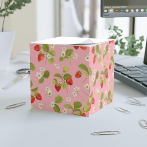 Könnte beinhalten: Ein quadratischer Notizblock in Rosa mit einem sich wiederholenden Muster aus roten Erdbeeren und weißen Blüten. Der Notizblock liegt auf einer weißen Oberfläche.