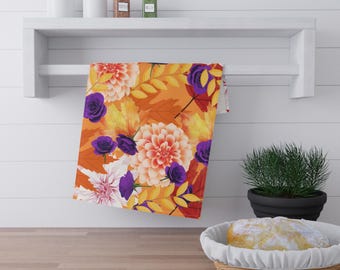 May include: A floral kitchen towel featuring a vibrant autumn design. The towel showcases large orange and white dahlias, purple roses, and yellow leaves against an orange background. The towel is hanging on a white shelf.