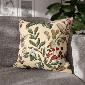 Op de afbeelding: Vierkant sierkussen met een crèmekleurige achtergrond met een botanische print. De print bevat groene bladeren, rode bessen en witte en bruine bloemelementen. Het kussen ligt op een grijze bank.