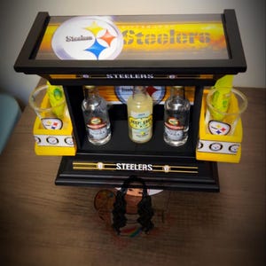 May include: A black and yellow Steelers-themed bar display with the team logo and the word "Steelers" prominently displayed. The display holds three small liquor bottles and two shot glasses. The background is a blurred image.